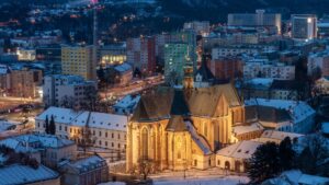 An evening view from above shows a church in the centre of the frame, with houses and other buildings beside it. Behind the church, the main road stretches through the neighbourhood as the light fades.