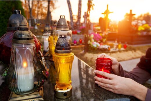 n the evening light, a person’s hands light one of several glass grave candles standing in sharp focus. Behind them, the cemetery fades into a soft blur, creating a calm and quiet atmosphere of remembrance.